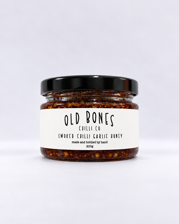 A jar of Old Bones Chilli Co Smoked Chilli Garlic Honey 200g, featuring a white label and a black lid, set against a plain light gray background.