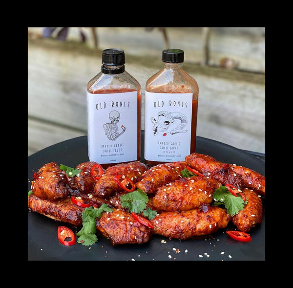 Two bottles of Old Bones Smoked Garlic Chilli Sauce with chicken wings on a black plate.
